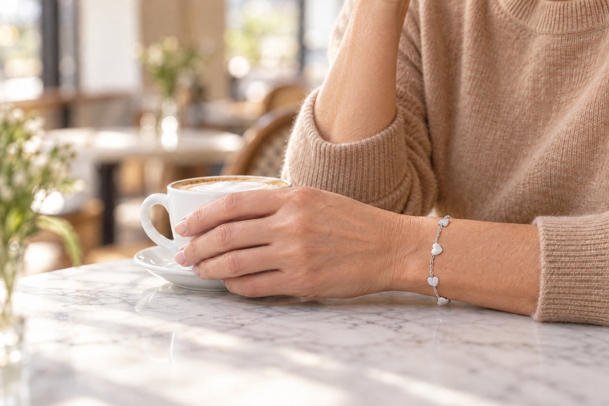 Frau im camelfarbenen Pullover hält im Café eine Tasse Cappuccino, am Handgelenk das silberne Armband mit Herzen